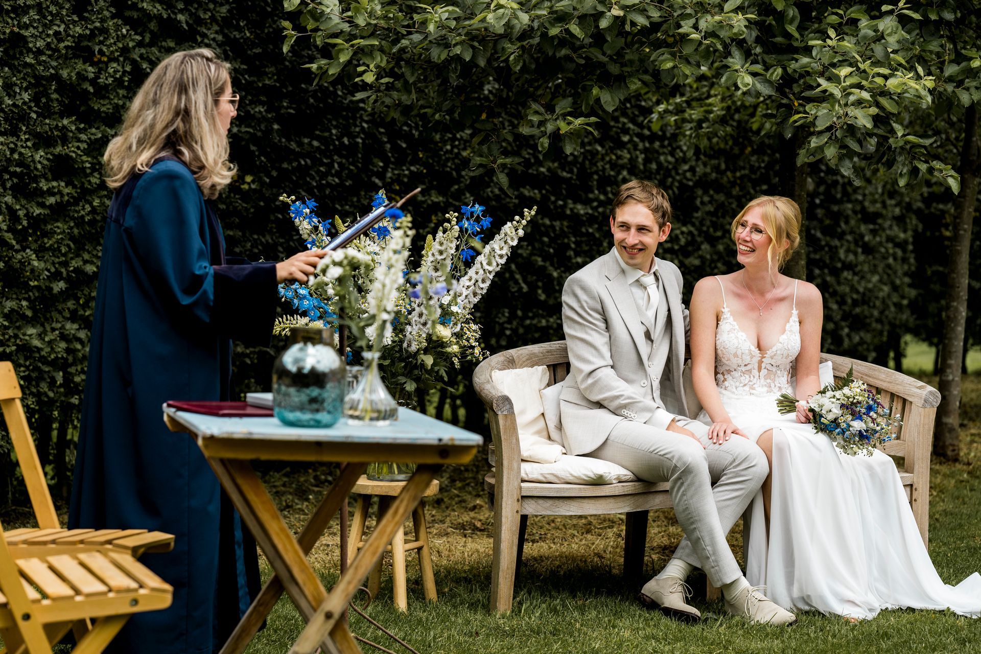Huwelijksceremonie: Bruid en bruidegroom zitten op een bankje, de ambtenaar van de burgerlijke stand in een blauwe toga staat erbij, bloemen. Buitenlocatie.