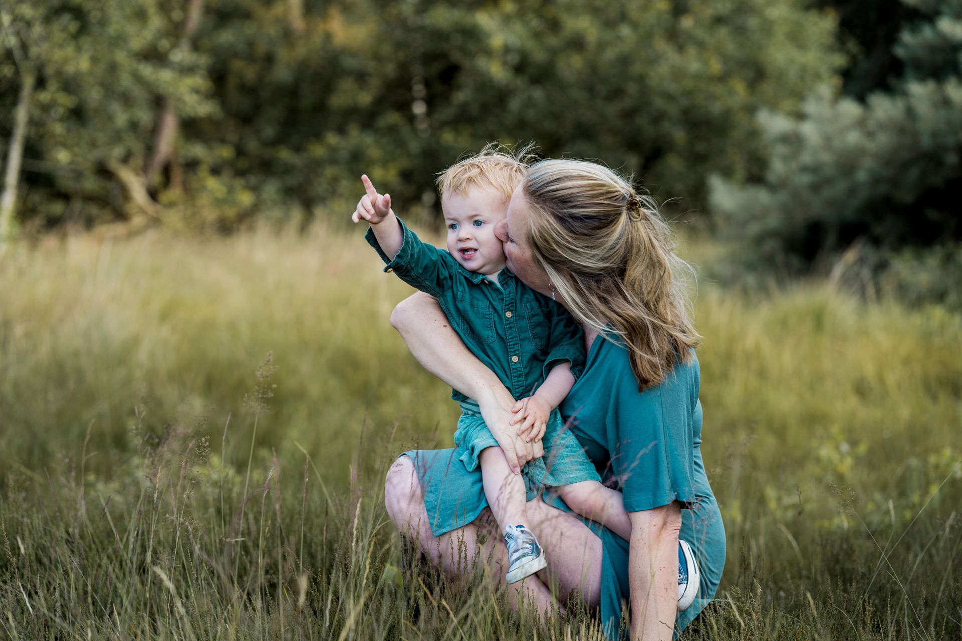Een vrouw knielt en houdt een kind vast. Het kind wijst omhoog. Ze staan ​​buiten in het hoge gras en dragen allebei turquoise kleding.