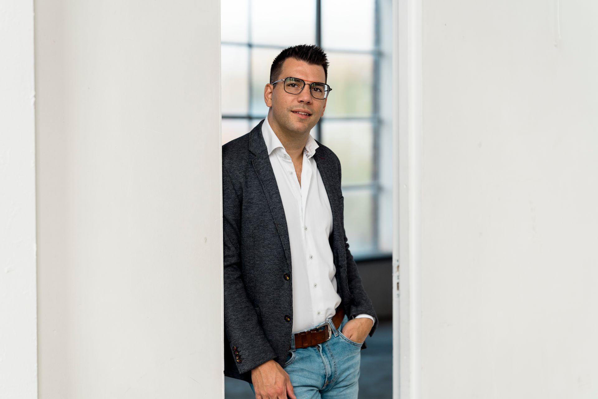 Een man in een blazer, spijkerbroek en bril staat in een deuropening, met zijn hand in zijn zak. Lichte, moderne omgeving.