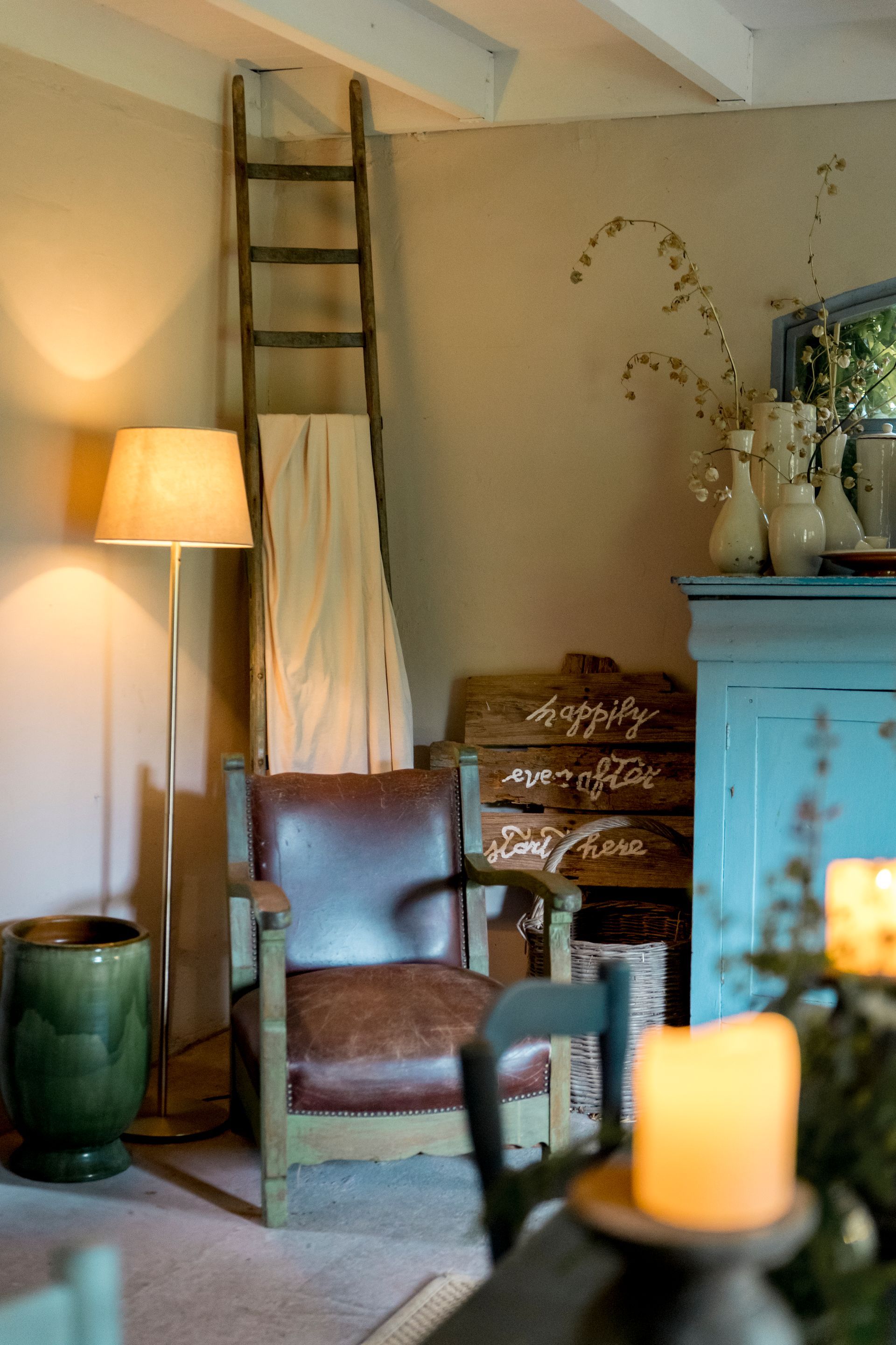 Gezellige zithoek met fauteuil, staande lamp, houten ladder, blauwe kast, kaarsen en vaas.