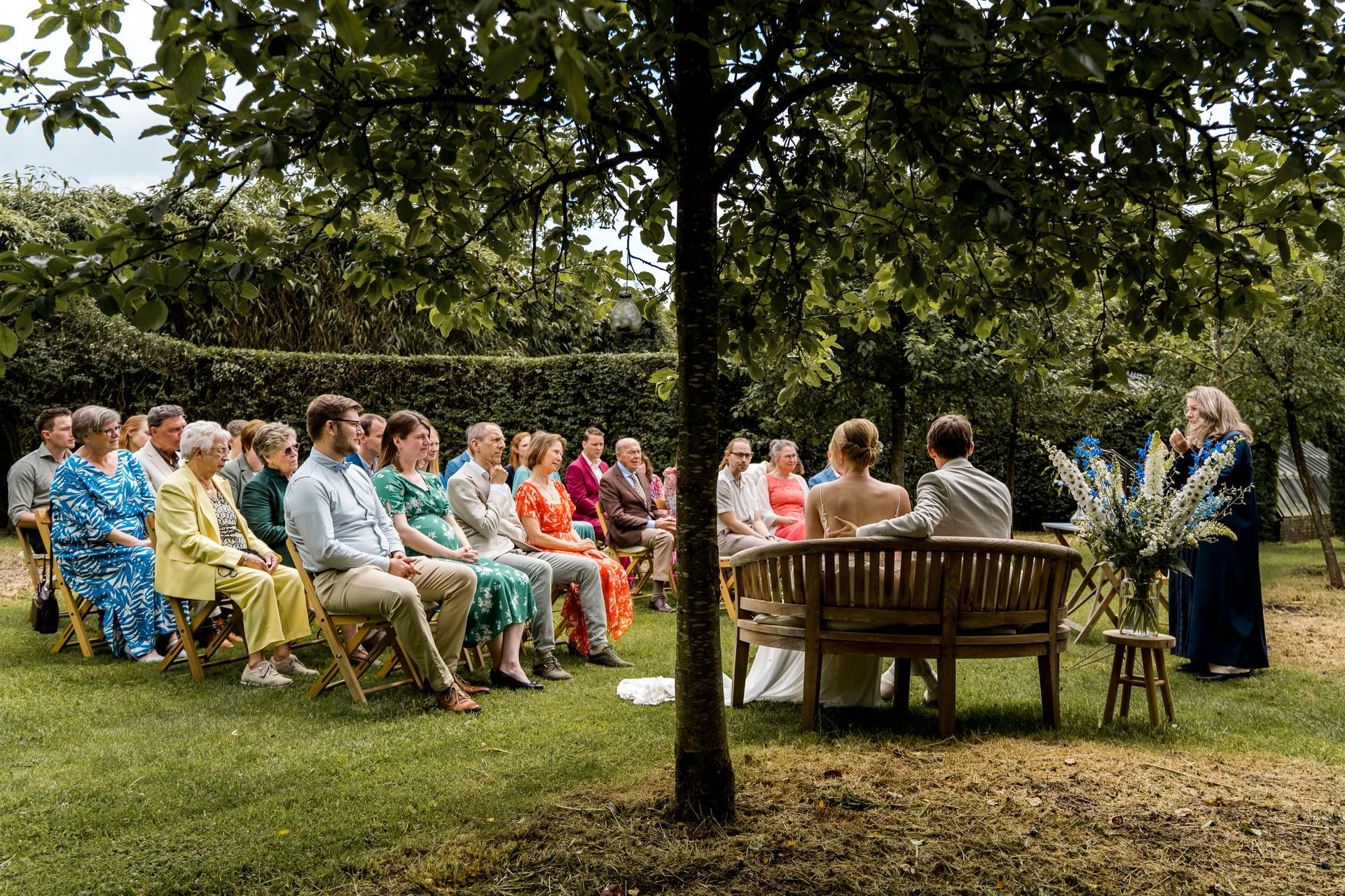 De huwelijksceremonie vindt buiten plaats, het bruidspaar zit, de gasten kijken toe en de ambtenaar van de burgerlijke stand spreekt onder een boom.