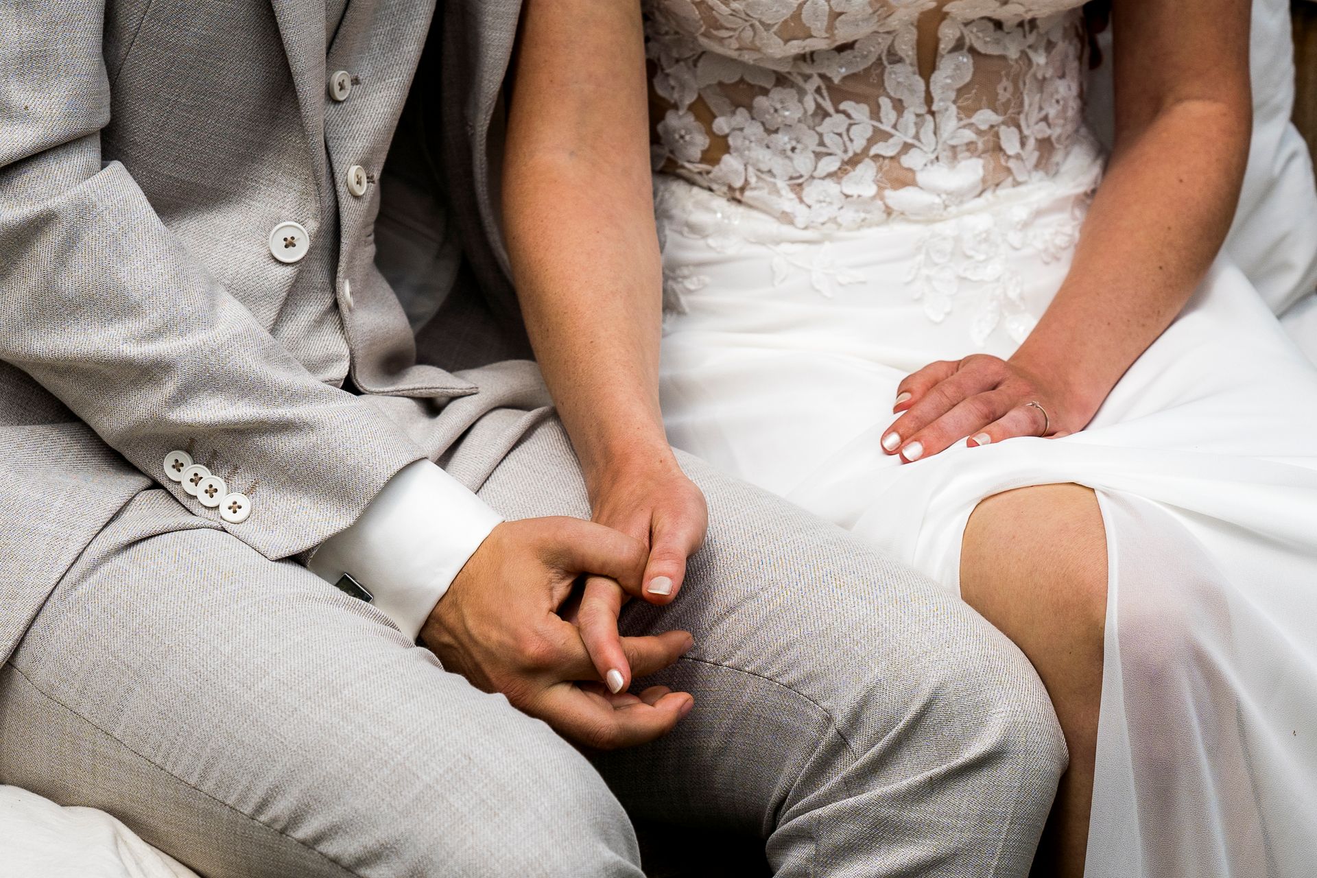 Een echtpaar, hand in hand, zit. De man draagt ​​een grijs pak en de vrouw een witte kanten jurk.