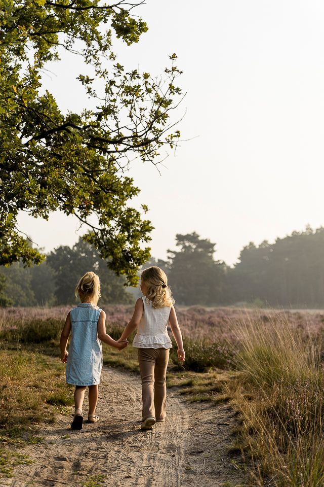 Twee jonge kinderen lopen hand in hand over een zandpad in een veld met paarse bloemen en bomen.