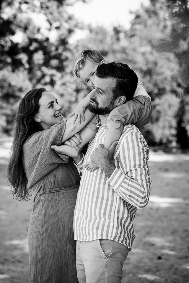 Familieportret: vader draagt kind op schouders, moeder lacht naar hen buiten. Zwart-wit.