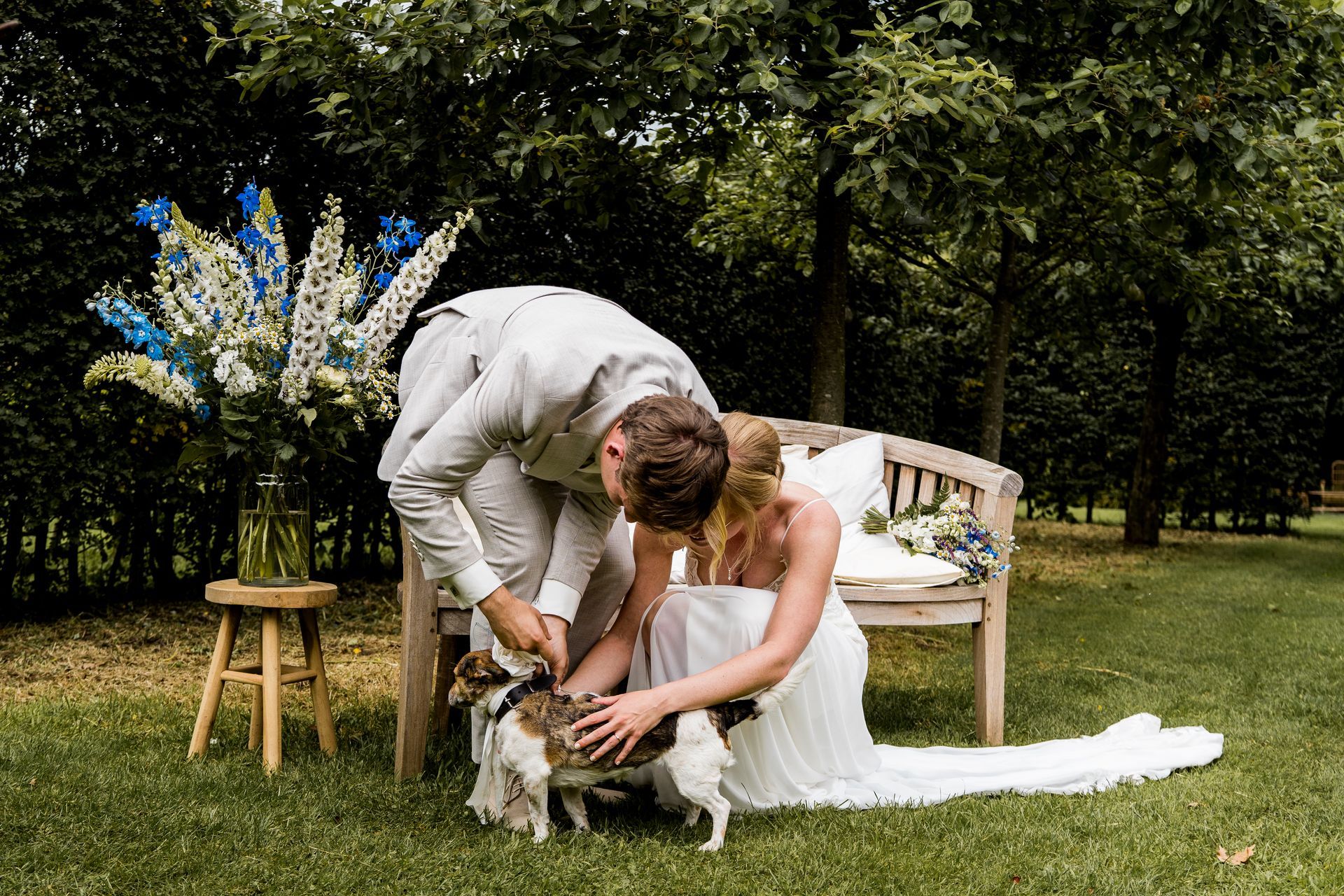 Een bruidspaar aait een hond in een tuin met bloemversieringen en een houten bankje.
