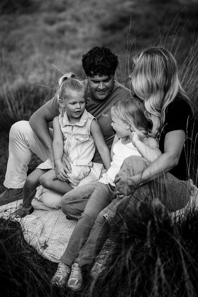 Een gezin van vier zit in het hoge gras. De kinderen kijken elkaar aan, de ouders kijken naar de kinderen. Zwart-wit.