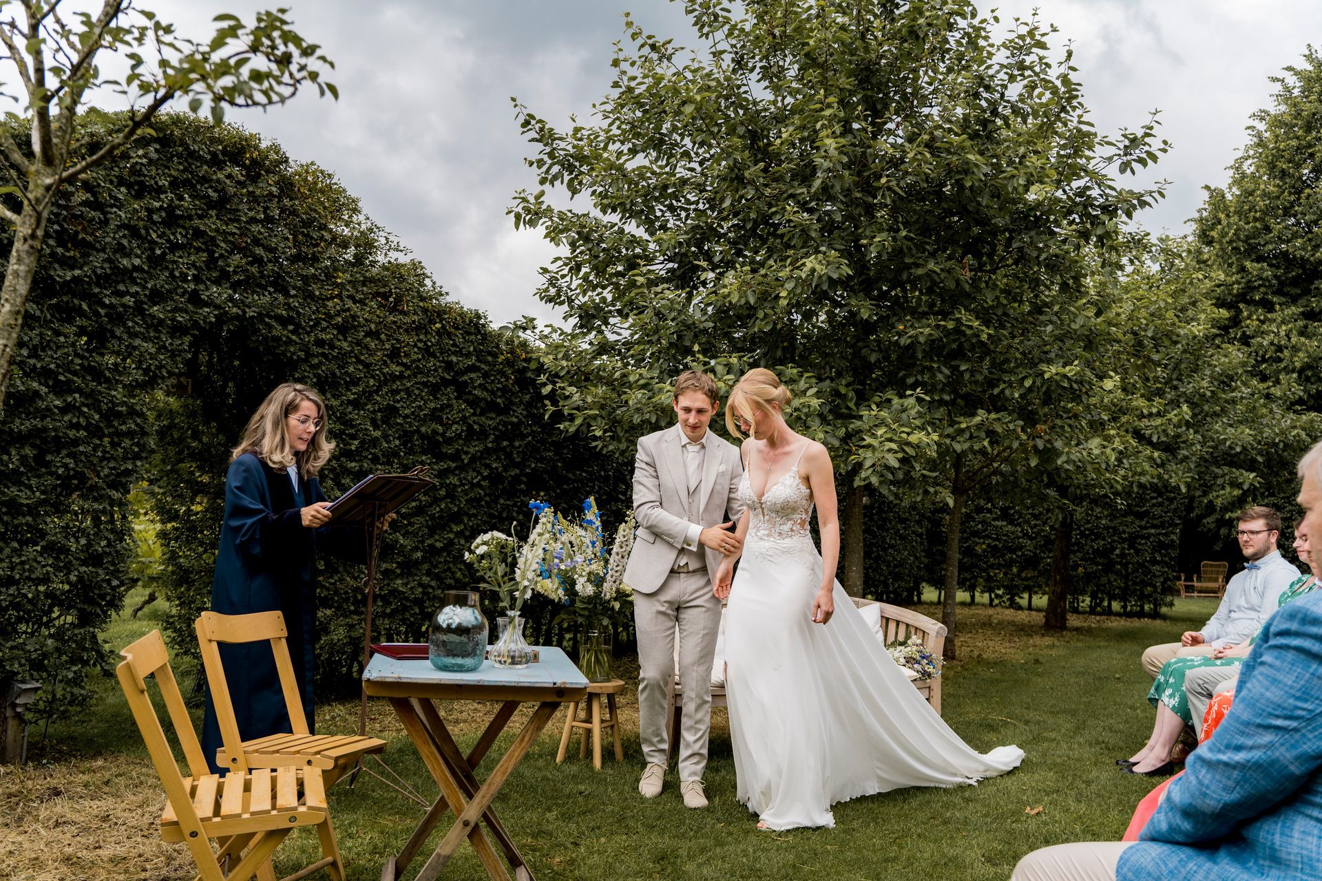 Twee mensen in trouwkleding houden elkaars hand vast tijdens een ceremonie in de buitenlucht. De ambtenaar van de burgerlijke stand en de gasten bevinden zich in een tuin.