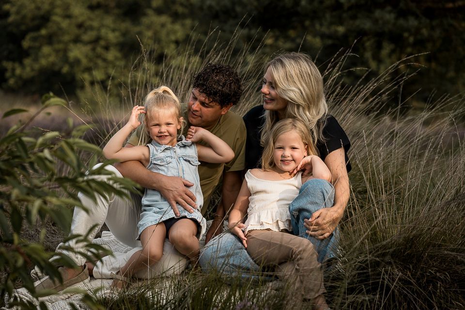 Een gezin van vier zit in het hoge gras; twee jonge meisjes, ouders met een glimlach.
