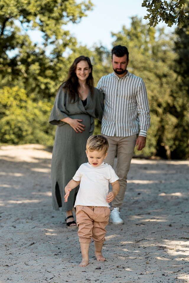 Een gezin in een park, moeder raakt de babybuik aan, vader glimlacht erachter, peuter loopt naar voren, zonnige dag.