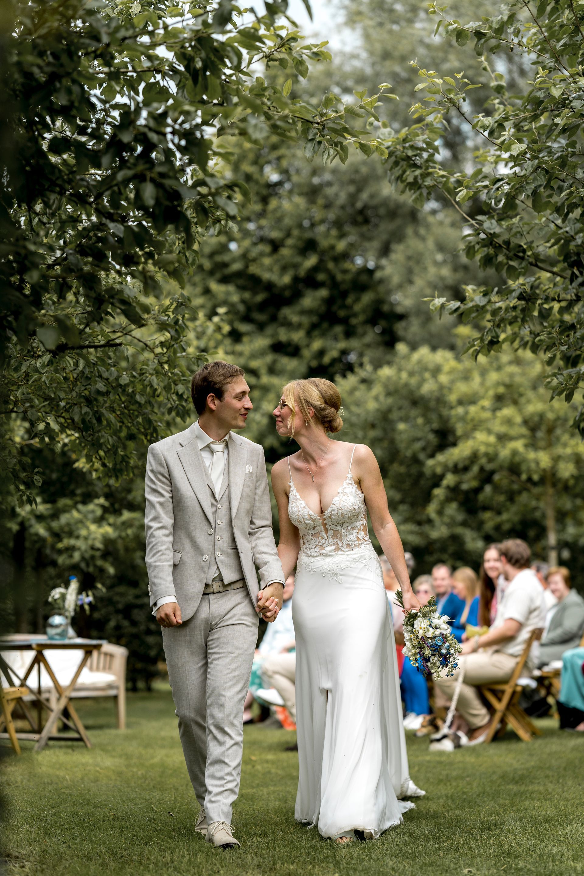 Het pasgetrouwde stel loopt hand in hand door een tuin na hun ceremonie. Ze kijken elkaar aan en glimlachen.