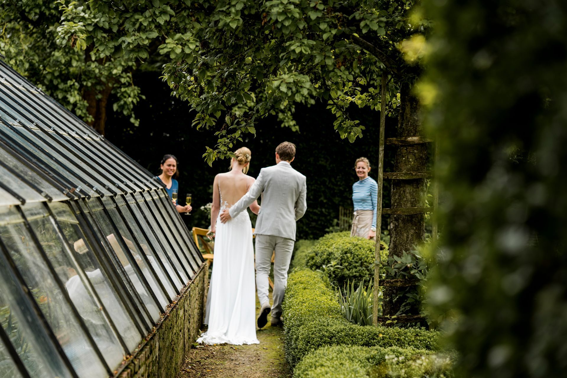 Bruid en bruidegom wandelen in een tuin; gasten kijken toe vanaf de kant van een glazen kas.