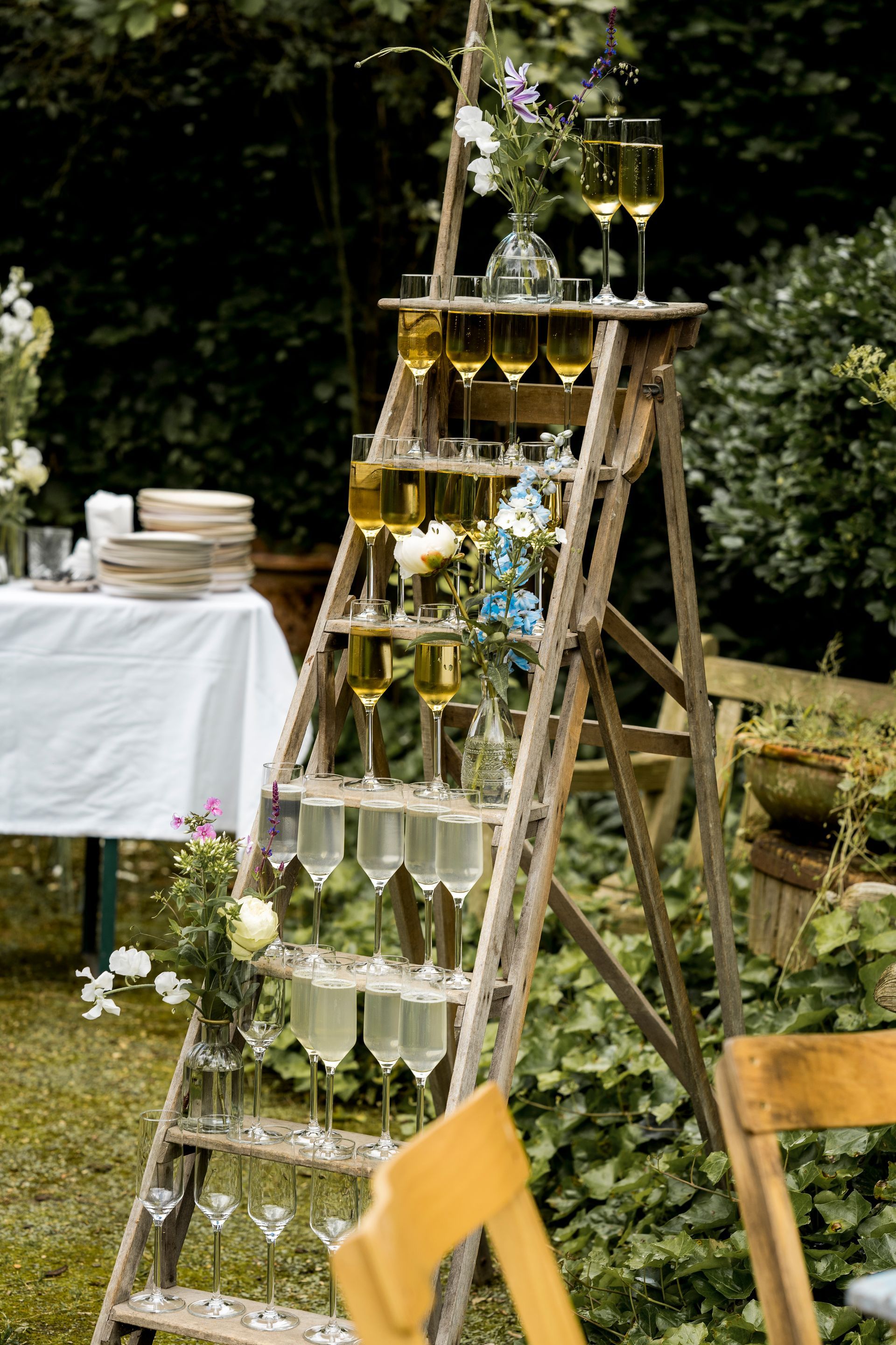 Een houten ladder met daarop vele champagneglazen staat buiten, versierd met bloemen.