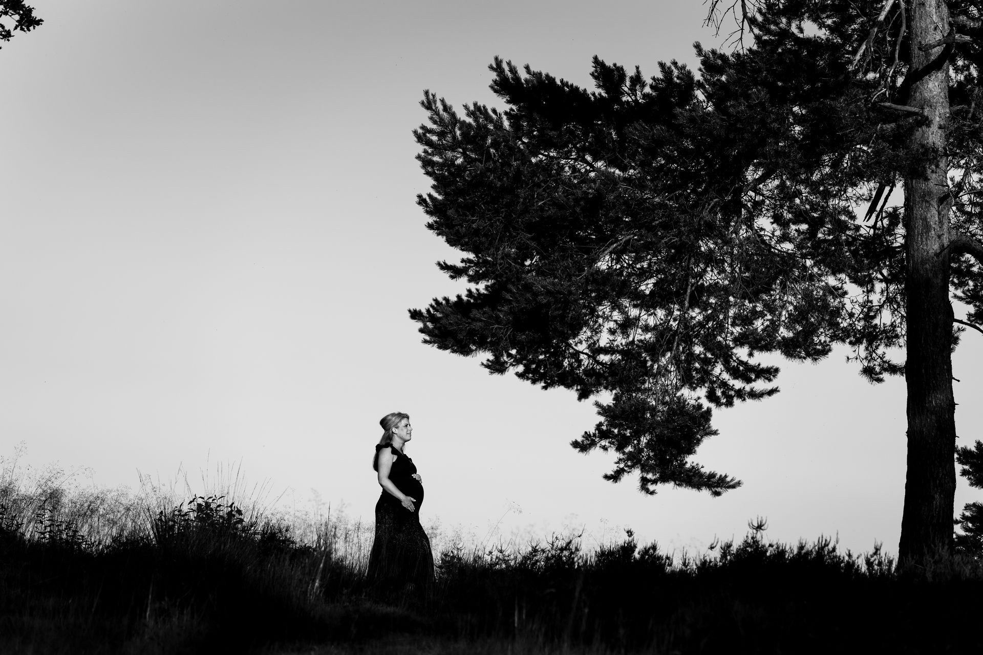 Een zwangere vrouw staat op een grasheuvel naast een grote boom, zwart-witfoto.
