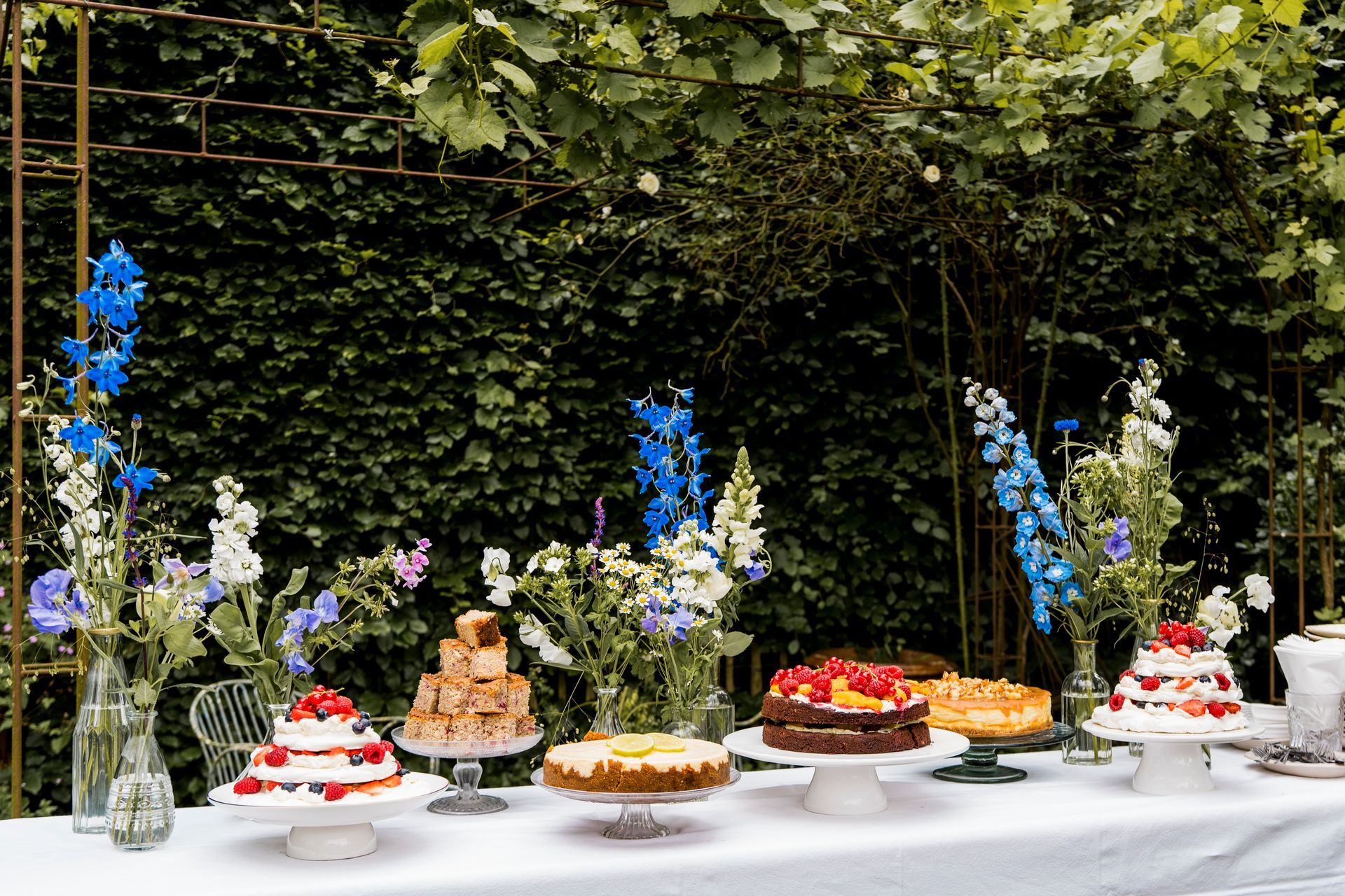 Een desserttafel in de buitenlucht met taarten, bloemen en een wit tafelkleed, tegen een groene muur van bladeren.