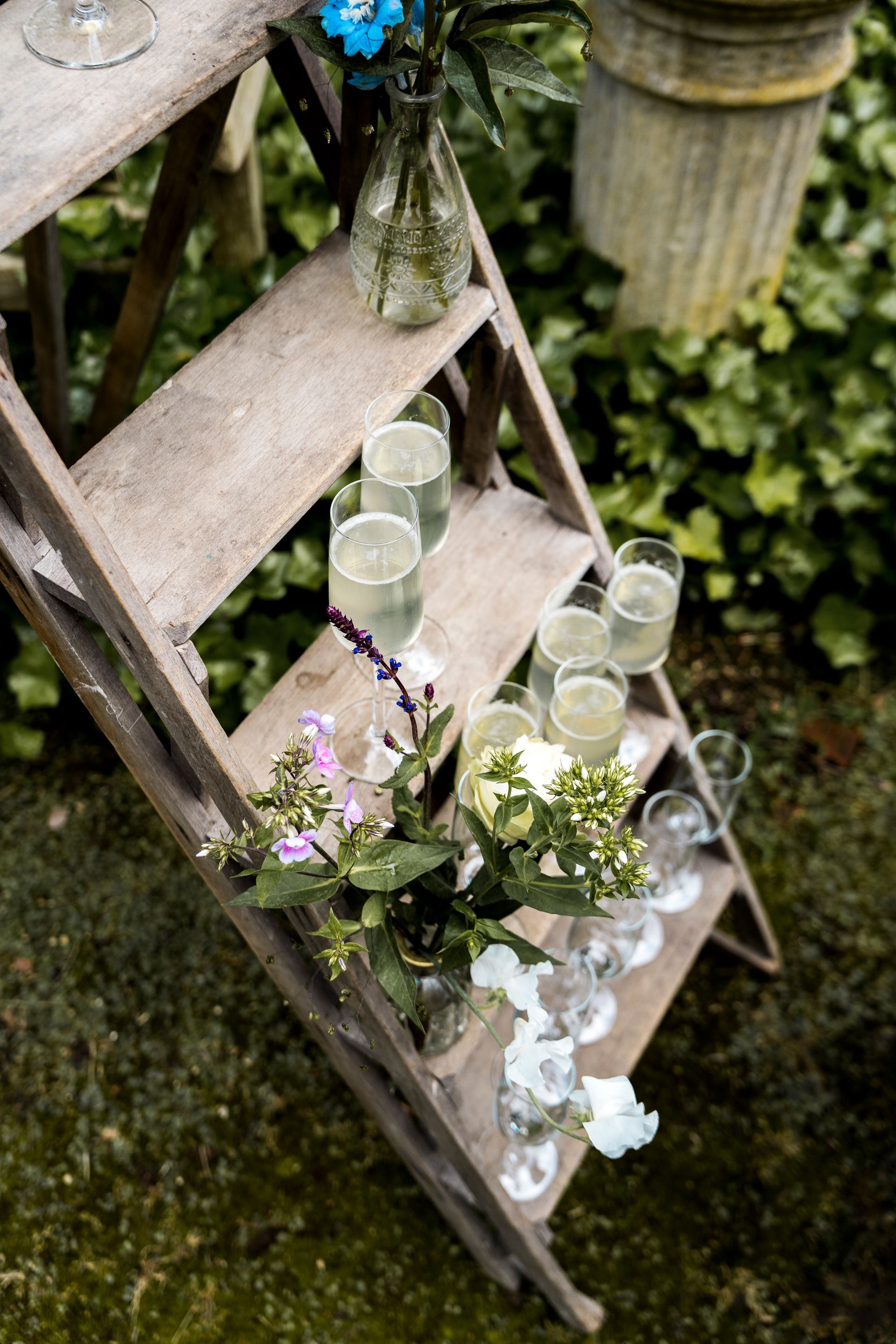 Een houten ladder met champagneglazen en bloemenvaasjes in een tuinsetting.