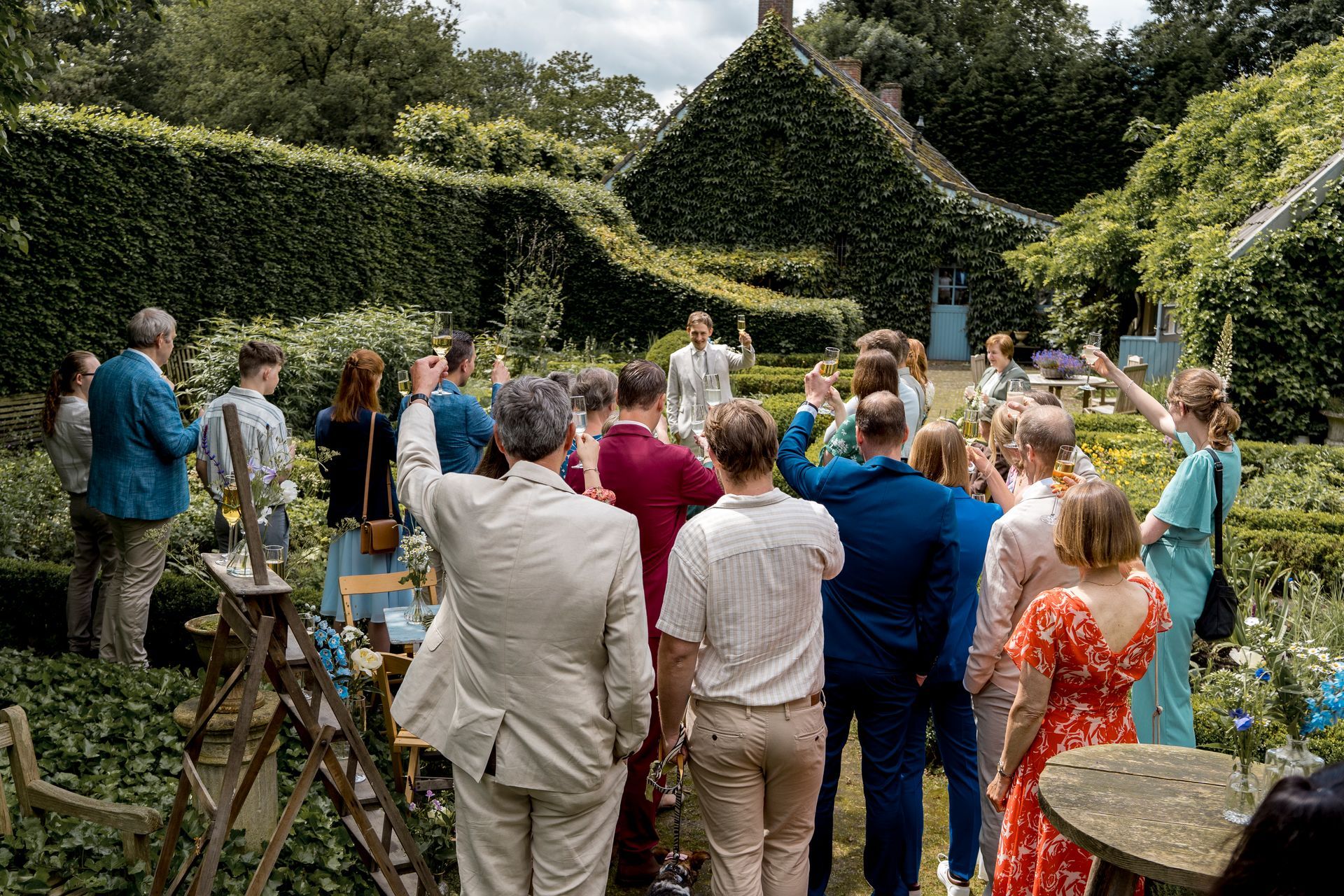 Een groep mensen brengt een toast uit in een tuin met een heg en een met klimop begroeid gebouw op de achtergrond.