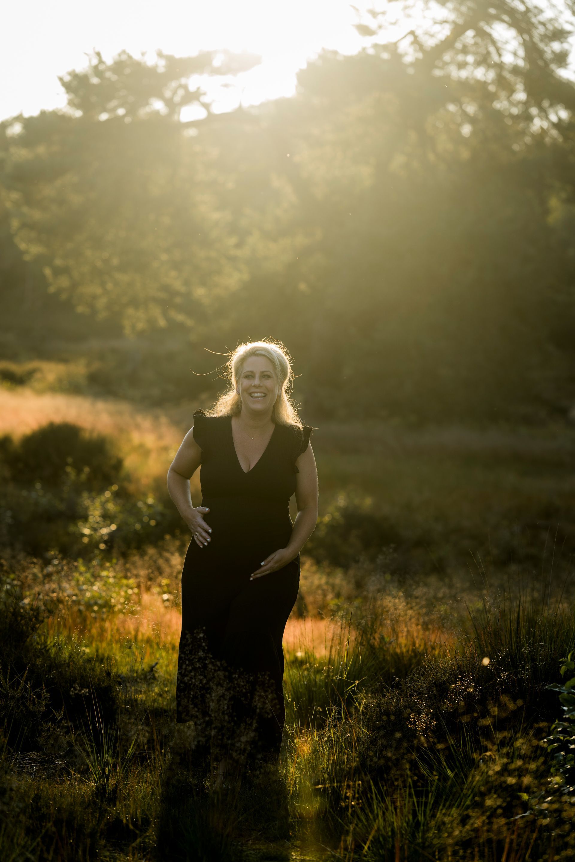 Een vrouw lacht in een veld, verlicht door de zon. Ze draagt ​​een donkere jurk en staat te midden van het hoge gras.