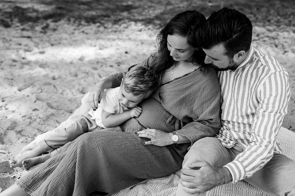 Familie op het strand: zwangere vrouw met kind en man, allen raken haar buik aan.