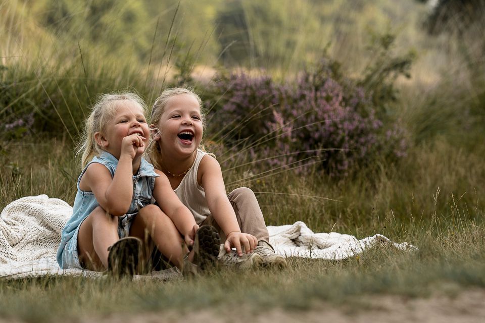 Twee jonge meisjes zitten lachend op een deken in een grasveld.