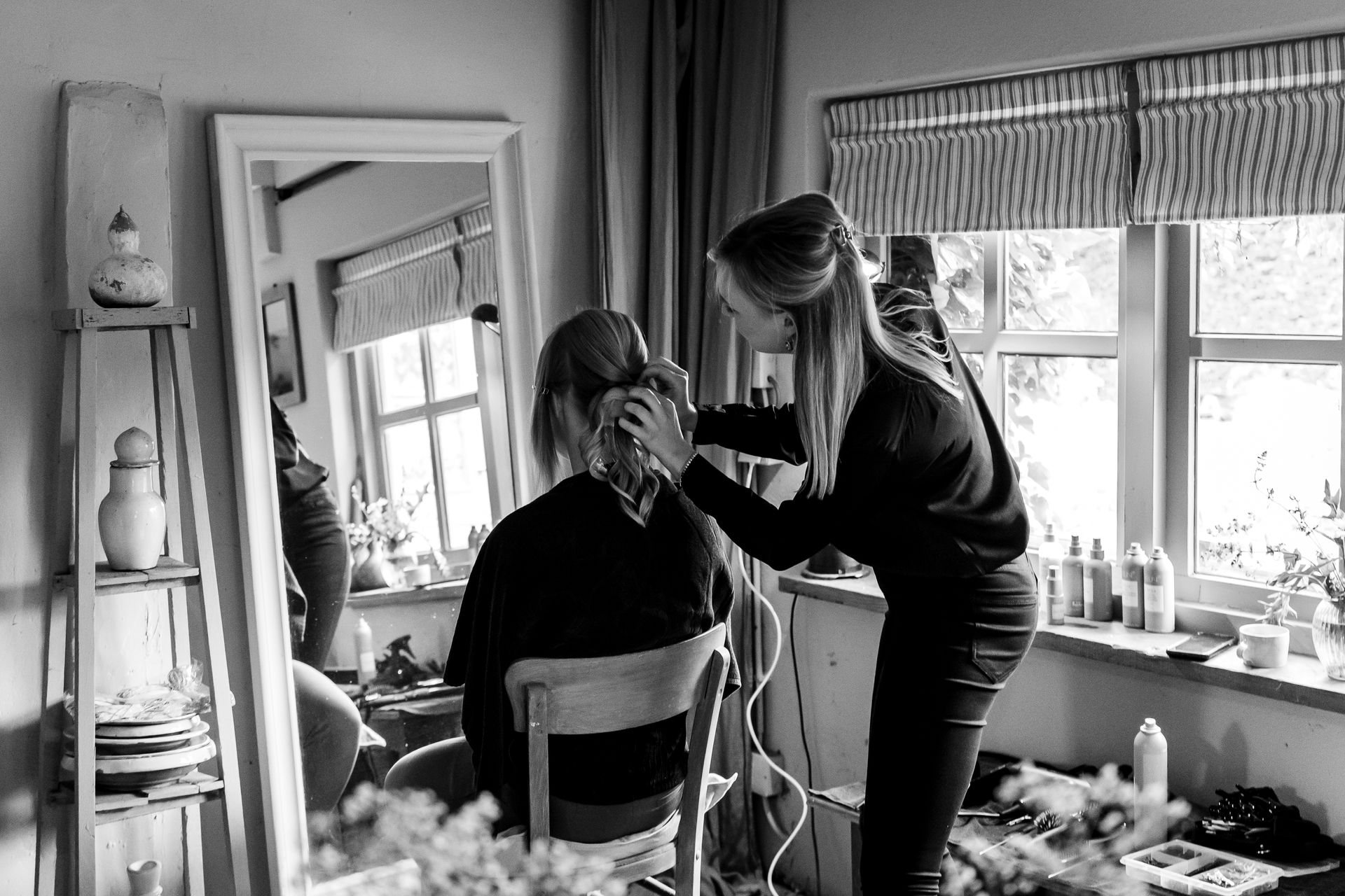 Een visagiste brengt make-up aan bij een persoon die voor een spiegel zit, verlicht door natuurlijk licht.