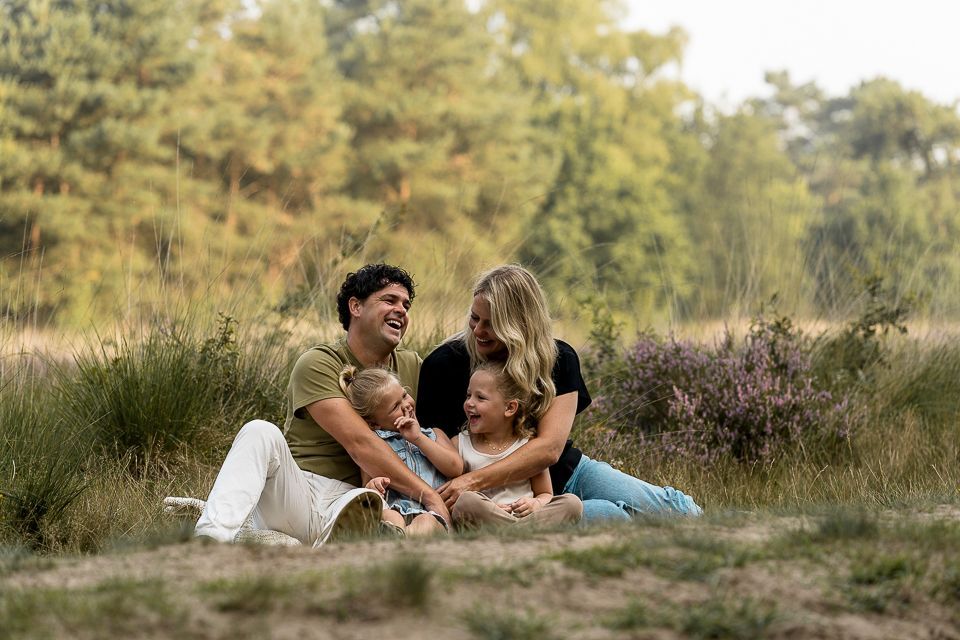 Een gezin van vier lacht in een grasveld. Twee jonge kinderen zitten bij hun ouders. Op de achtergrond is een bos te zien.