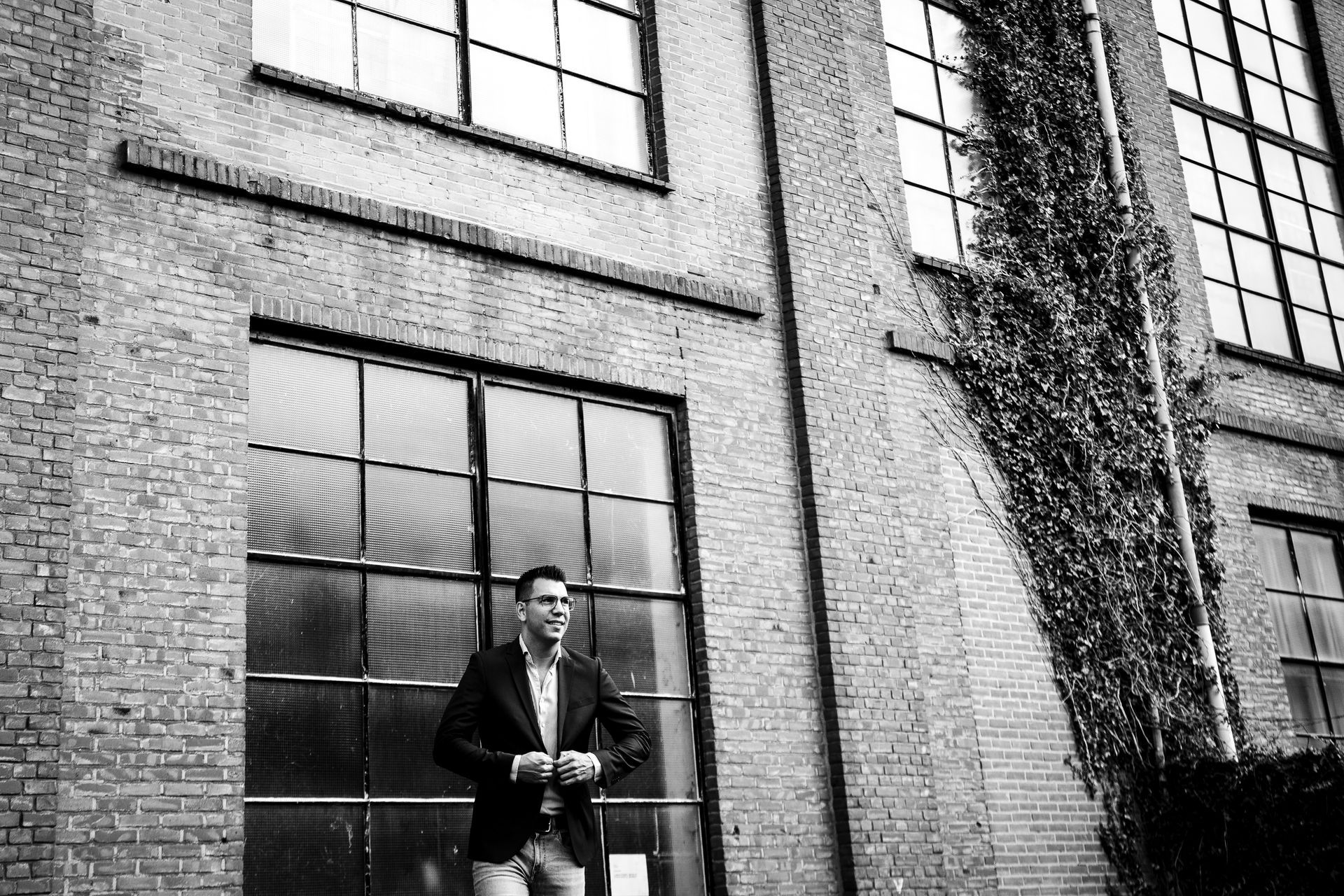 Een man in een blazer staat bij een bakstenen gebouw met grote ramen.