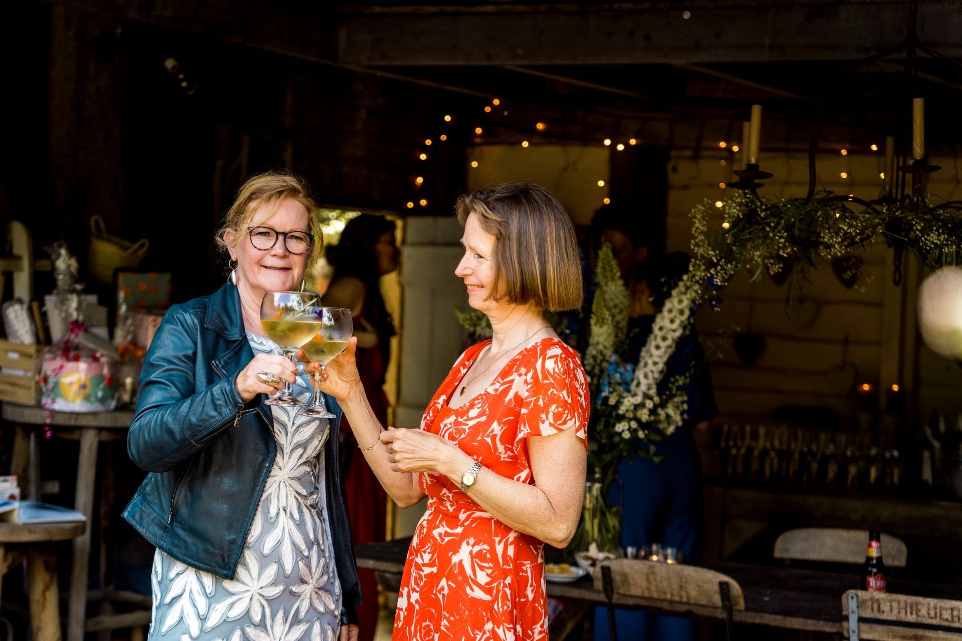 Twee vrouwen klinken hun glazen en glimlachen. De ene draagt ​​een rode jurk, de andere een zwart jasje en een jurk met een patroon, tijdens een bijeenkomst.