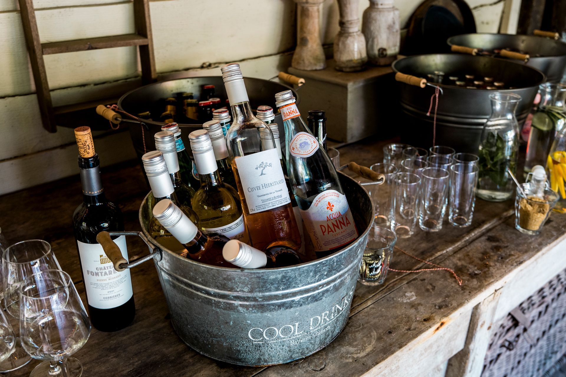 Flessen wijn en andere dranken in metalen bakken, uitgestald op een rustieke houten bar.