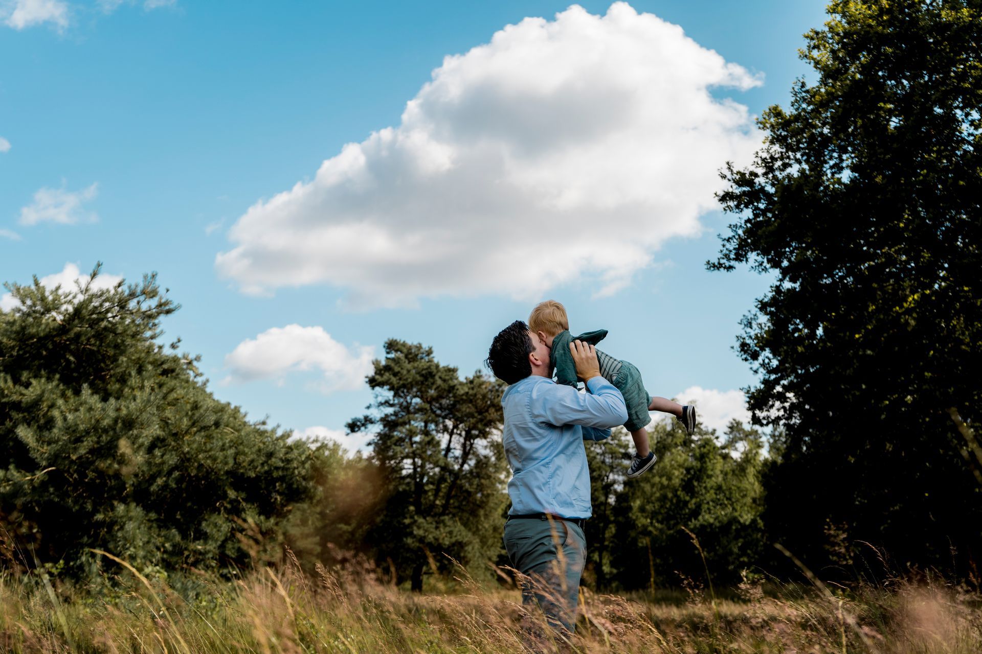 Een man tilt een kind hoog in de lucht onder een gedeeltelijk bewolkte hemel. Ze bevinden zich in een grasveld, omringd door bomen.