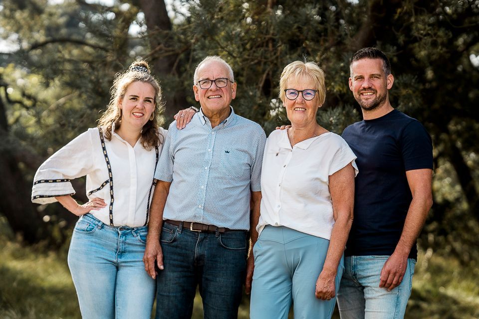 Een gezin poseert buiten voor een portret: een ouder echtpaar, twee volwassenen, lachend. Groene bomen op de achtergrond.