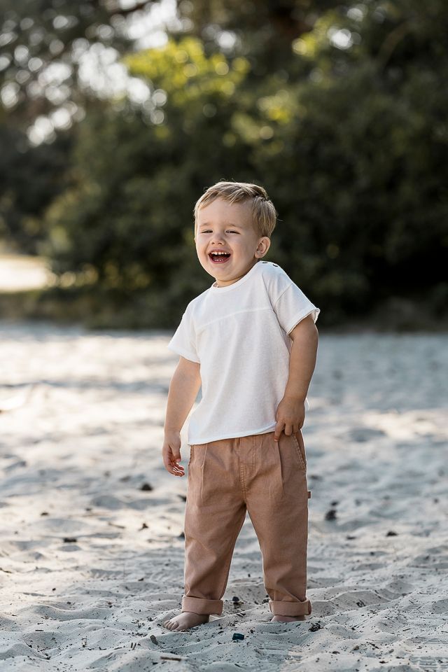 Een jong kind lacht, staand in het zand, gekleed in een wit shirt en een bruine broek, met wazige groene bomen op de achtergrond.