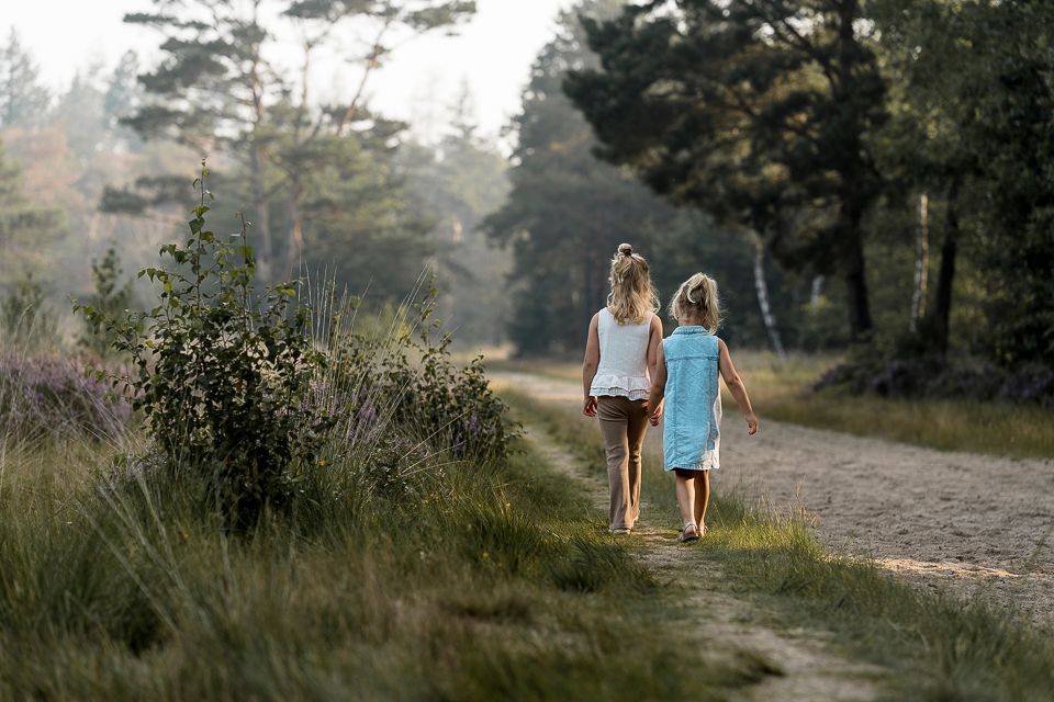 Twee kinderen lopen over een zandpad in een bos; zonlicht filtert door de bomen.