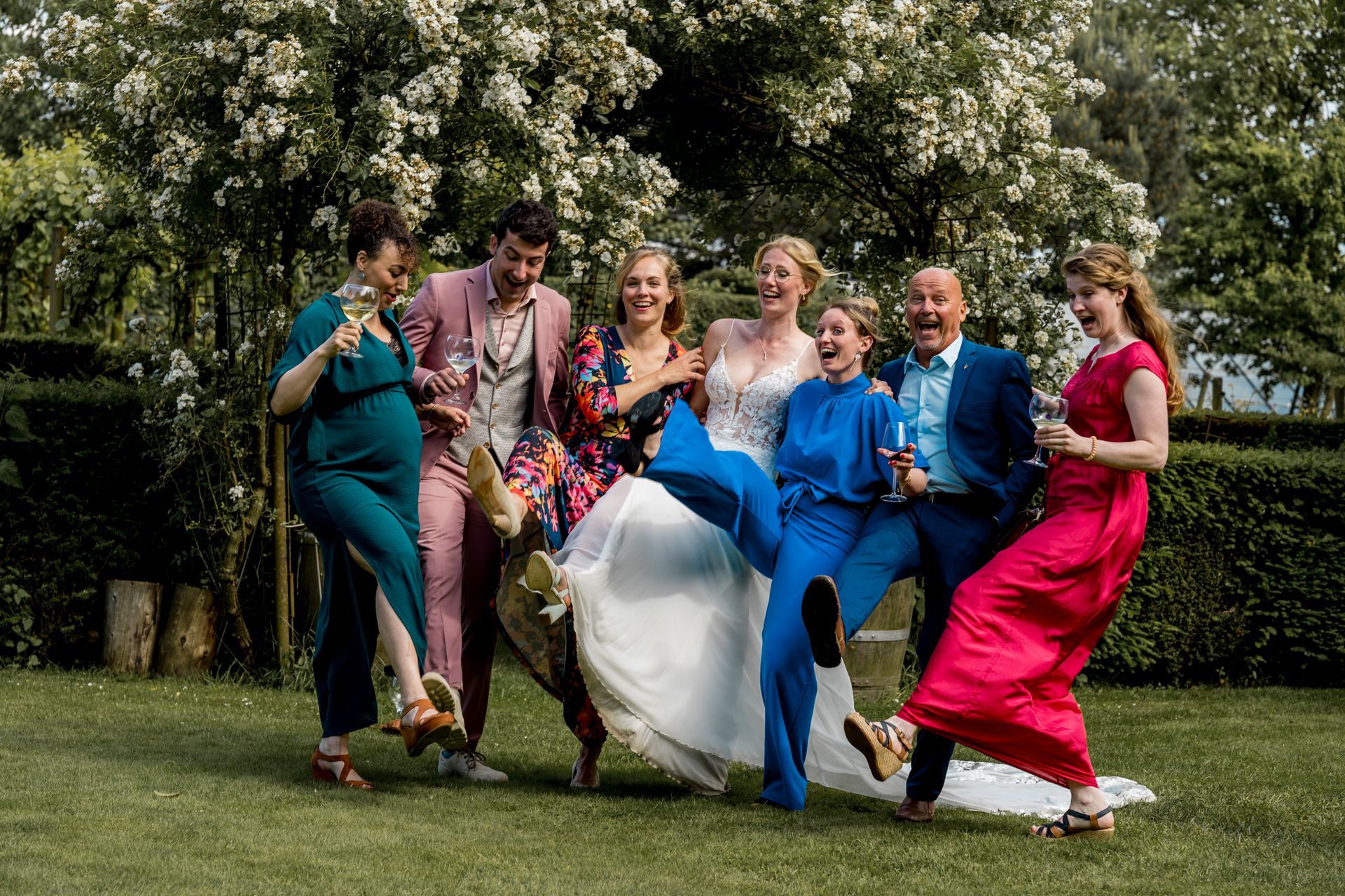 Een groep bruiloftsgasten poseert speels in de buitenlucht. Ze dragen opvallende kleding: blauw, roze, rood en bloemenprints. De bruid staat in het midden.