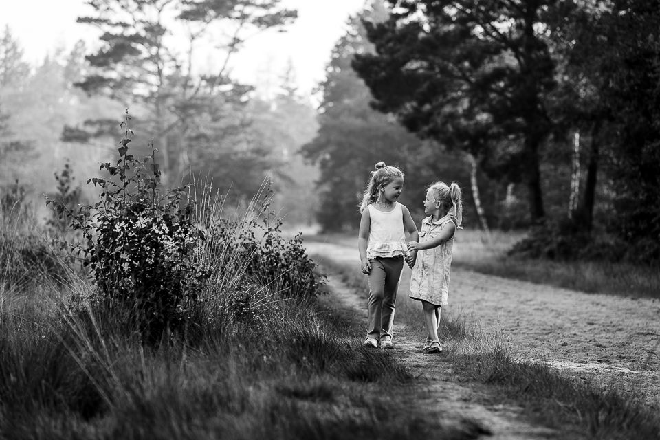 Twee jonge meisjes, hand in hand, lopen over een pad in een bos. Zwart-wit.