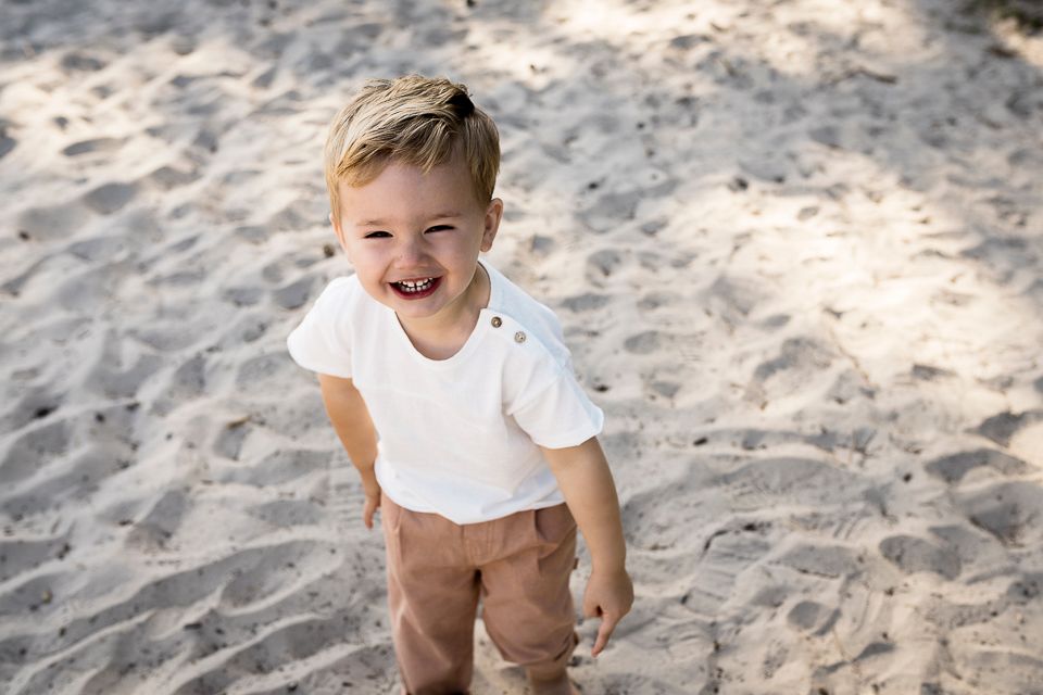Een vrolijk kind staat in het zand, gekleed in een wit shirt en een bruine broek, en lacht.