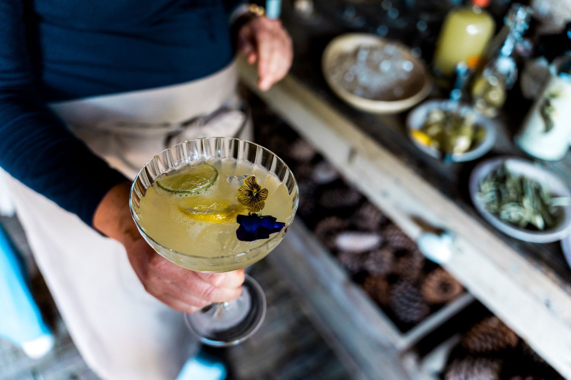 Een persoon houdt een cocktail vast, gegarneerd met limoen en bloemen, bij ingrediënten op een houten tafel.