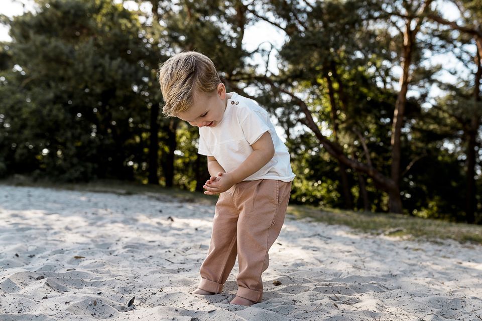 Blond peutertje in een wit shirt en beige broek, kijkt naar beneden terwijl ze in het zand staat, met bomen op de achtergrond.