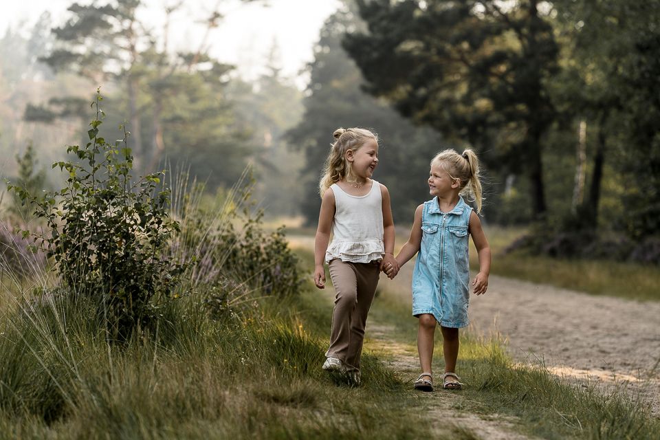 Twee jonge meisjes lopen hand in hand over een zandpad in een bos en glimlachen.