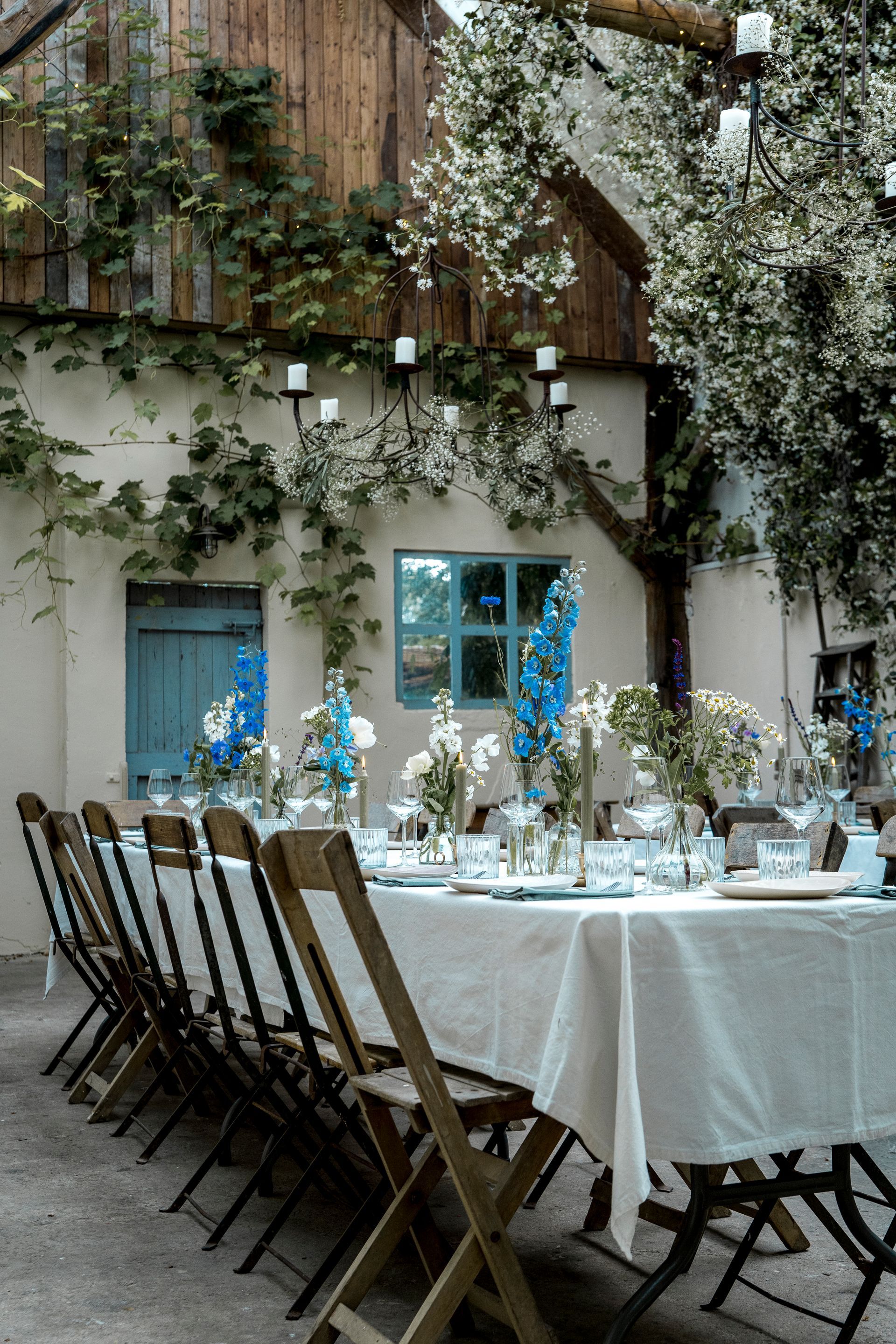 Een eettafel in de tuin, gedekt voor een maaltijd en versierd met blauw-witte bloemen, staat naast een gebouw dat begroeid is met klimplanten.