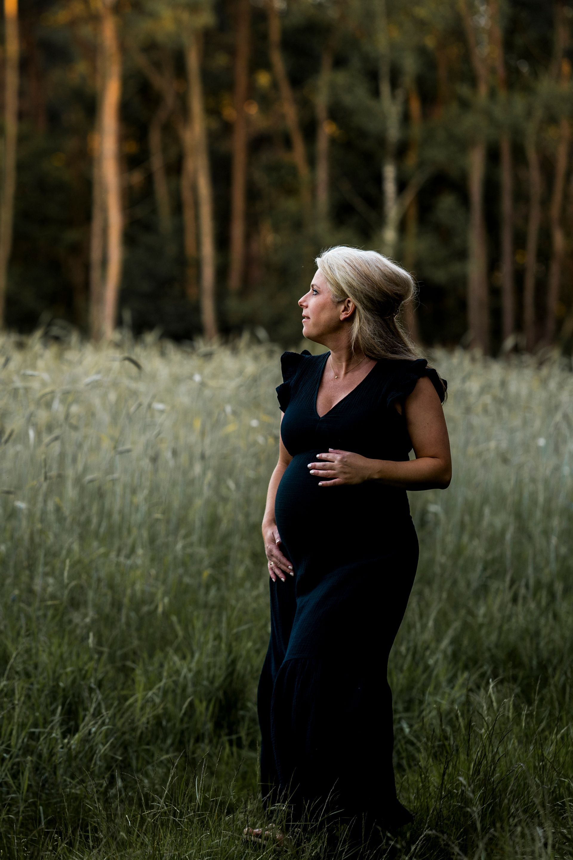 Een zwangere vrouw in een zwarte jurk, met haar hand op haar buik, staat in een veld met hoog gras en bomen op de achtergrond.