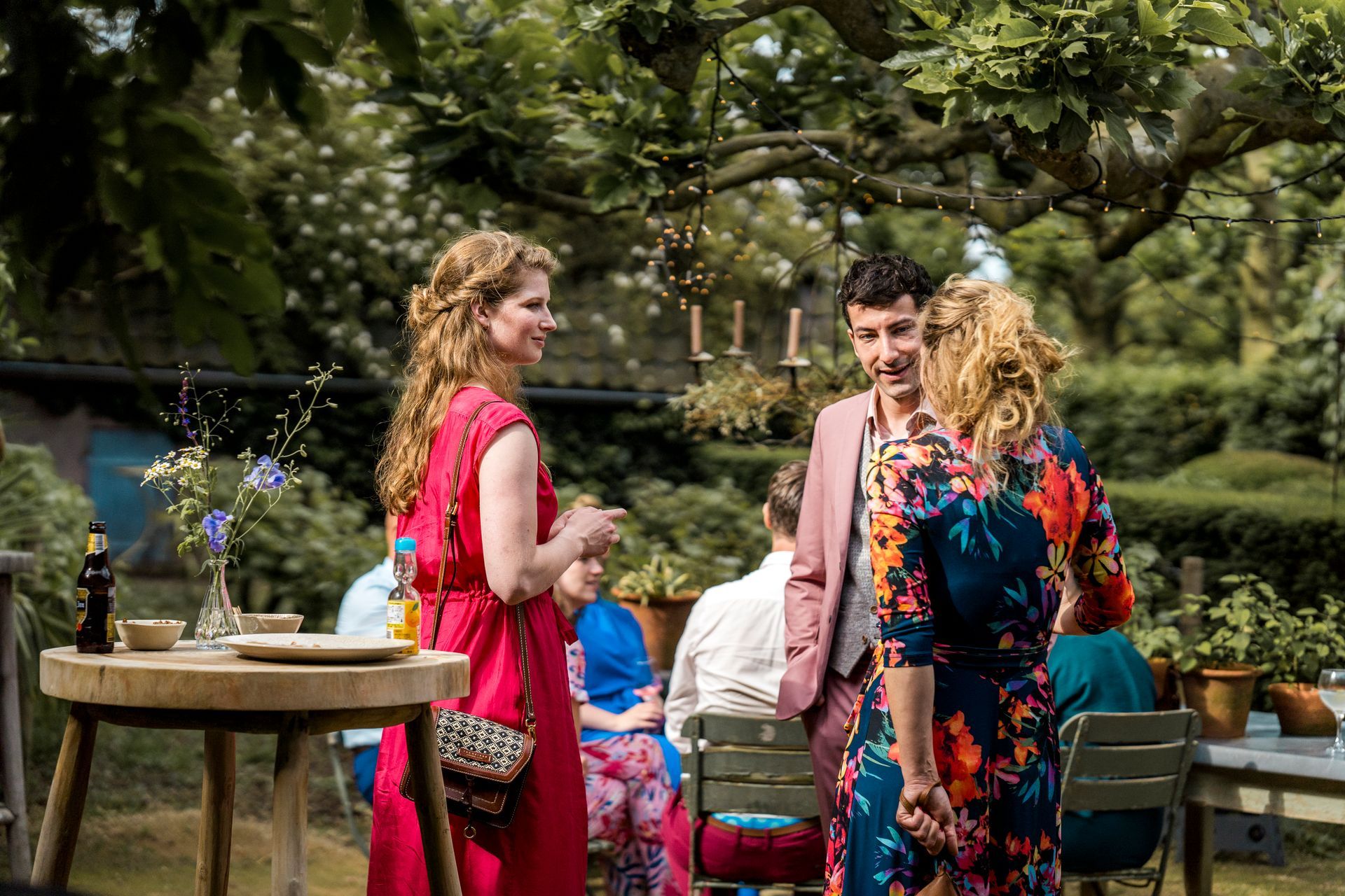 Mensen in gesprek tijdens een bijeenkomst in de buitenlucht. Een vrouw in rood lacht, een man in roze en een vrouw in een jurk met bloemenprint.