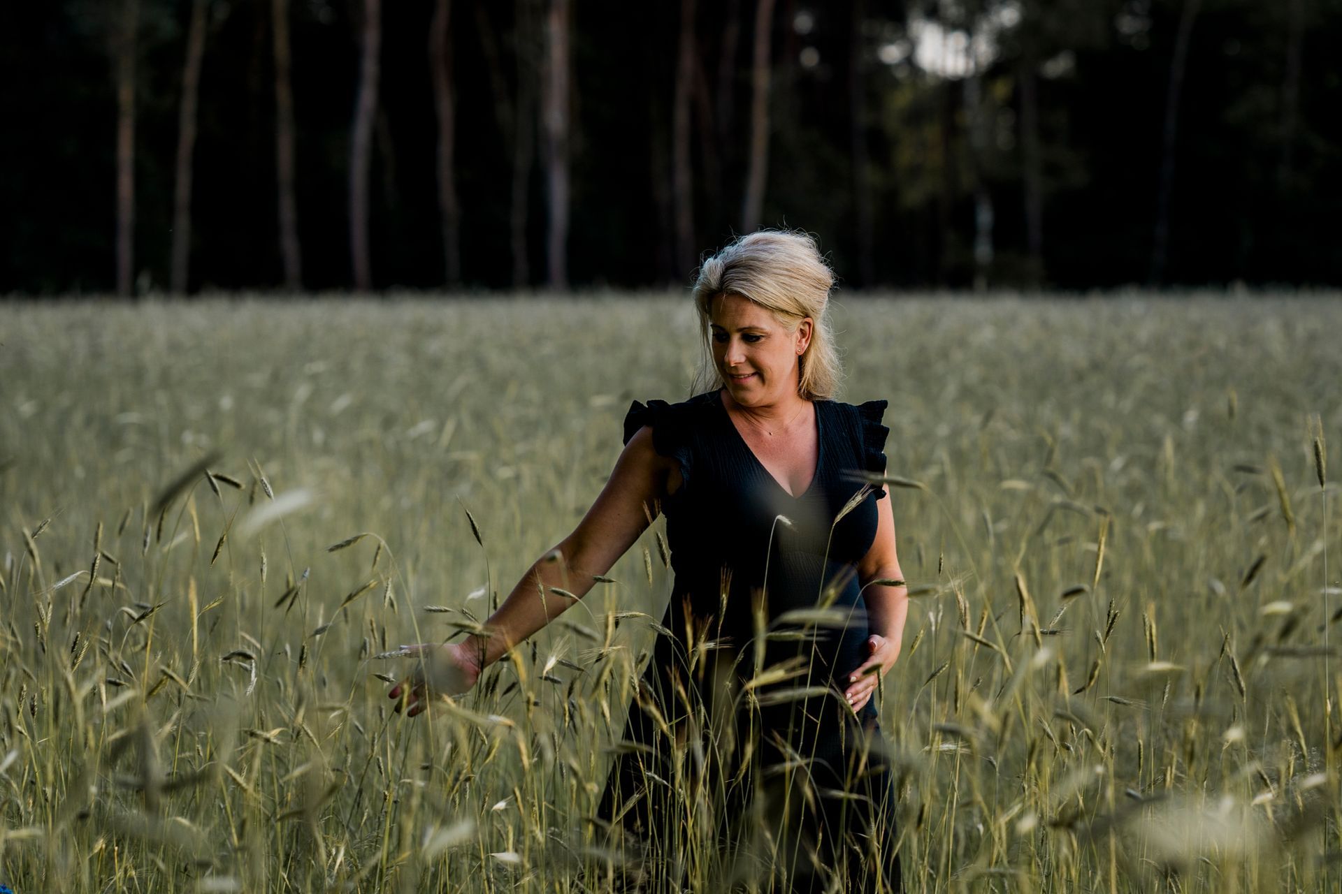 Vrouw in zwarte jurk staat in een veld met hoog gras, glimlacht en raakt de planten aan.