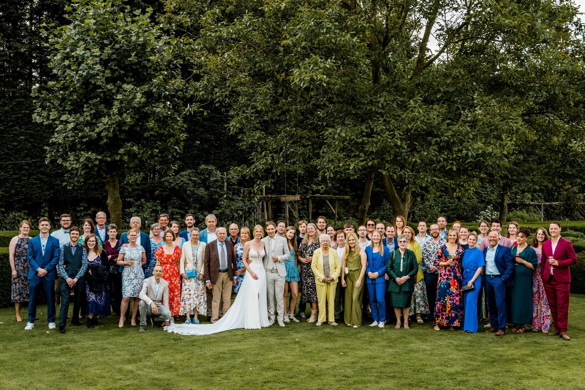 Een grote groep mensen poseert voor een foto op een gazon voor bomen; een huwelijksfeest.