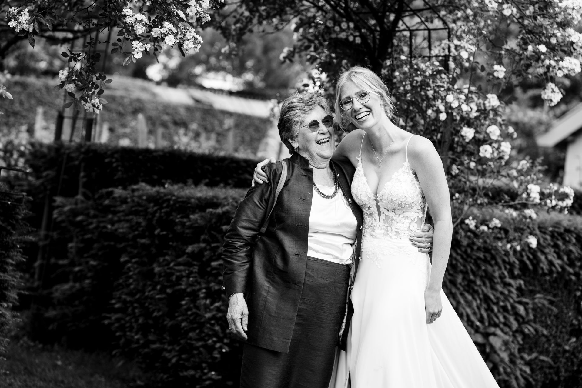 Bruid in witte jurk omhelst oudere vrouw buiten onder bloemen. Beiden glimlachen.
