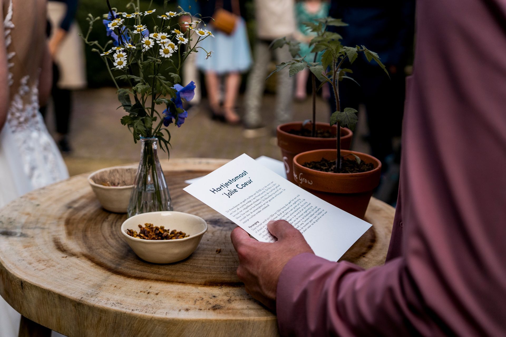 Een persoon met een vel papier in zijn hand staat naast potplanten en bloemen op een houten tafel.