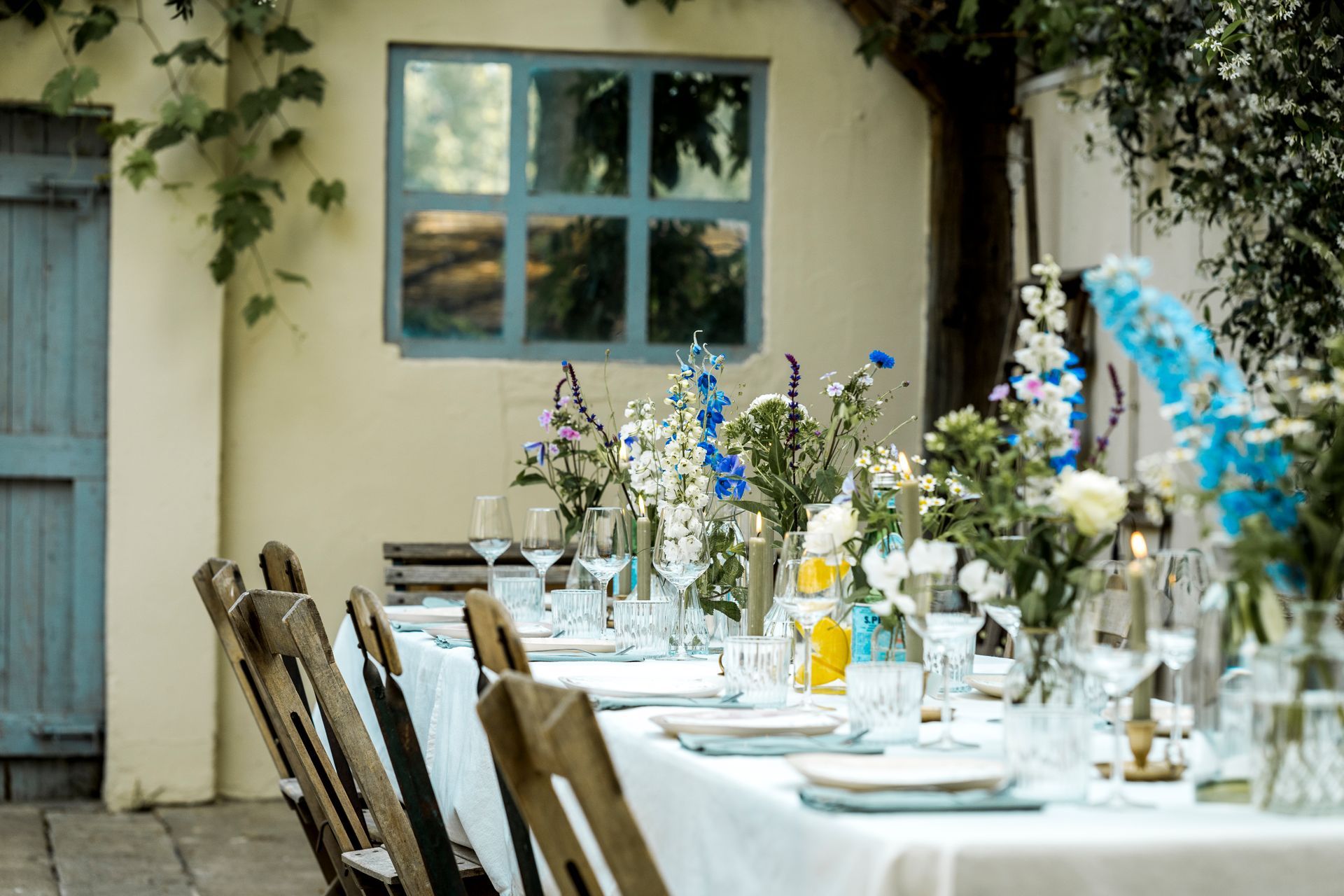 Een gedekte eettafel voor buiten, geschikt voor een feestelijke gelegenheid, met een wit tafelkleed, blauwe accenten en bloemstukken in het midden.