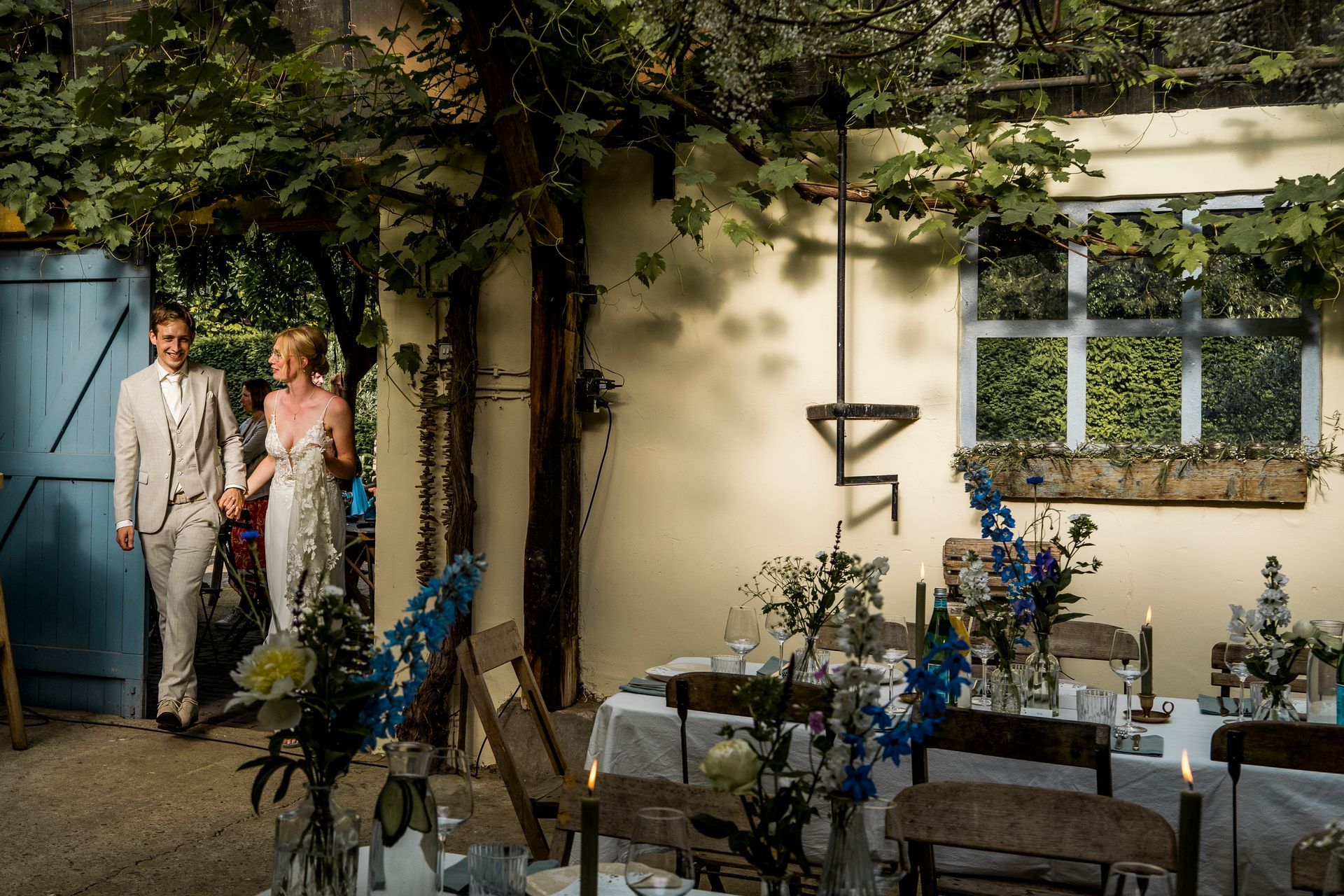 Bruid en bruidegroom verlaten de blauwe deur en betreden de buitenruimte voor de huwelijksreceptie. De tafels zijn gedekt met bloemen en kaarsen.