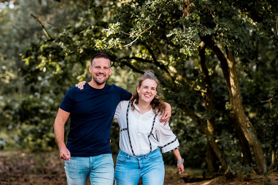 Een man en een vrouw glimlachen, met hun armen om elkaar heen geslagen, terwijl ze buiten langs bomen wandelen.