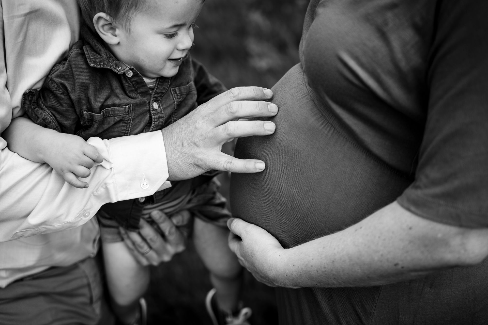 Een kind raakt de buik van een zwangere vrouw aan; een andere persoon houdt het kind vast. Zwart-witfoto.