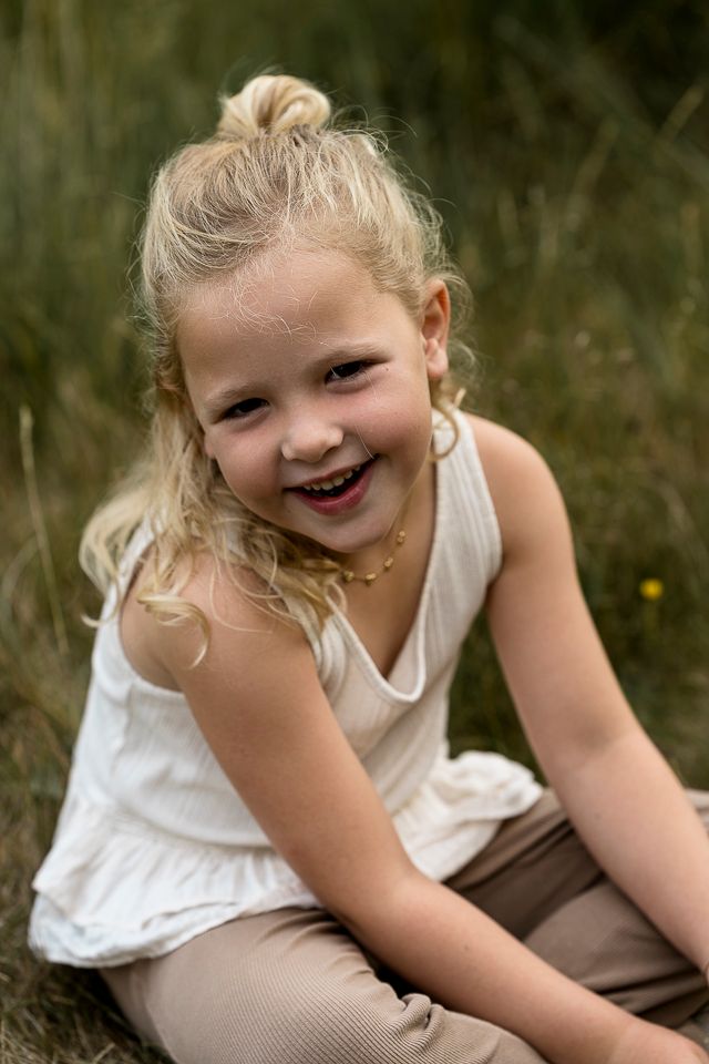 Blond kindje zit lachend buiten in het gras, gekleed in een wit topje en een bruine broek.